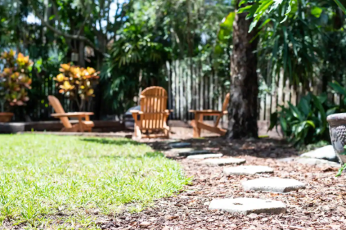 A serene garden with stone pathways, lush greenery, and wooden chairs nestled among trees and plants.