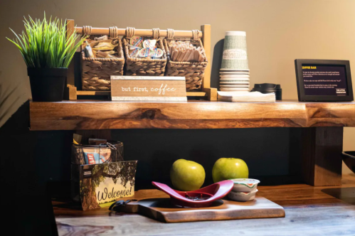 A wooden shelf with baskets of snacks, coffee cups, green apples, and a sign that says "but first, coffee."