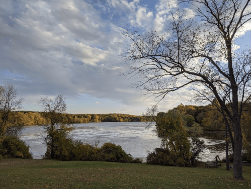 A serene river scene with autumn foliage, trees, and a cloudy sky reflecting on the water's surface.