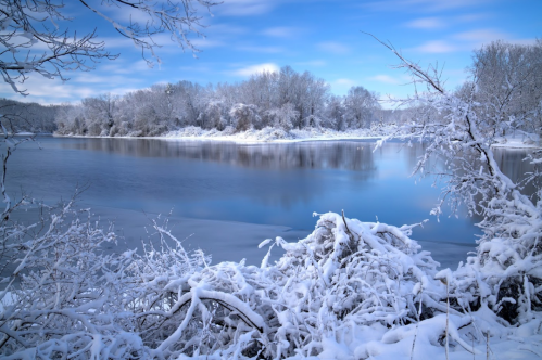 A serene winter landscape featuring a snow-covered riverbank and trees under a clear blue sky.