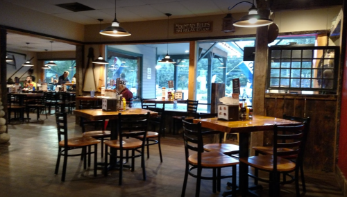 Interior of a rustic restaurant with wooden tables, chairs, and large windows, creating a cozy dining atmosphere.
