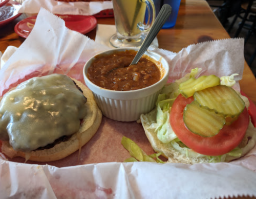 A cheeseburger with melted cheese, lettuce, tomato, and pickles, served with a side of chili in a white bowl.