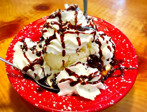 A red plate with a scoop of vanilla ice cream topped with whipped cream and chocolate syrup, garnished with powdered sugar.