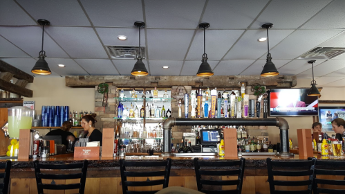 A bar with shelves of liquor, a bartender, and patrons seated at tables in a casual dining setting.
