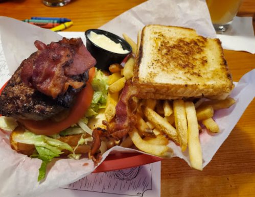 A burger topped with bacon, lettuce, and tomato, served with fries and a side of dipping sauce on a wooden table.