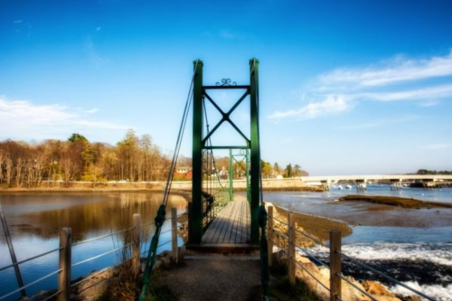 A green suspension bridge over a calm river, surrounded by trees and a clear blue sky.