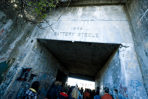 A group of people walking through a large, weathered archway marked "1342 BATTERY STEELE," surrounded by graffiti.