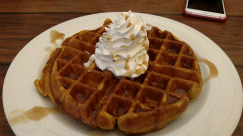 A golden waffle topped with whipped cream and drizzled with syrup, served on a white plate.