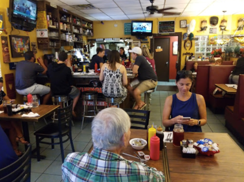 A busy restaurant interior with patrons at the bar and tables, featuring colorful decor and a TV in the background.
