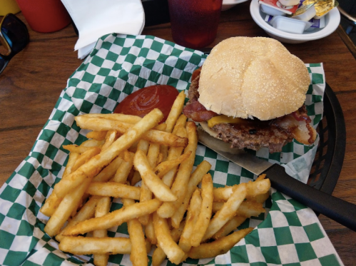 A burger with bacon and cheese on a sesame seed bun, served with a side of golden fries and ketchup in a checkered basket.