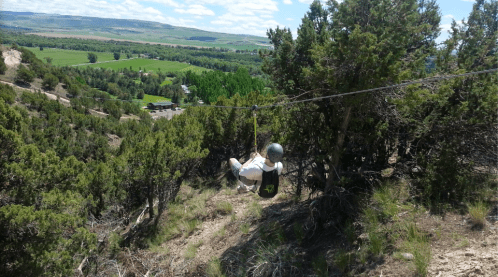 A person zip-lining over a green landscape with trees and hills under a blue sky with clouds.