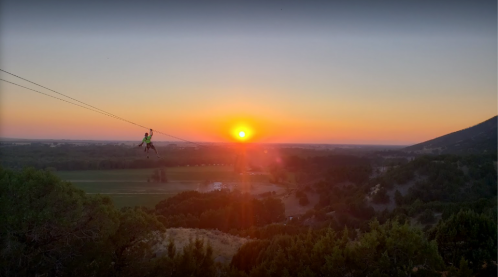 A person zip-lining against a vibrant sunset over a scenic landscape with hills and fields.