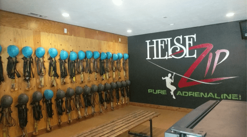 A wall of blue helmets and harnesses in a zipline facility, with a sign reading "HEISE Zip - Pure Adrenaline!"