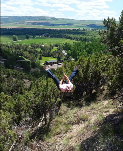 A person hangs upside down on a zip line, surrounded by green trees and a scenic valley below.