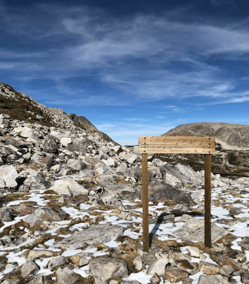 A wooden sign stands on rocky terrain with snow patches, under a blue sky with wispy clouds.