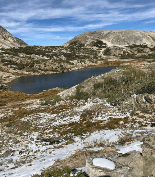A serene mountain landscape featuring a calm lake surrounded by rocky terrain and patches of snow under a blue sky.