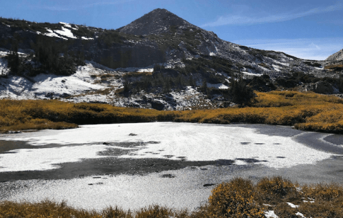 A serene mountain landscape with a partially frozen lake, surrounded by autumn foliage and snow-capped peaks.