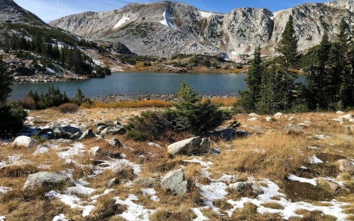A serene mountain landscape featuring a lake surrounded by rocky terrain, trees, and snow-capped peaks in the background.