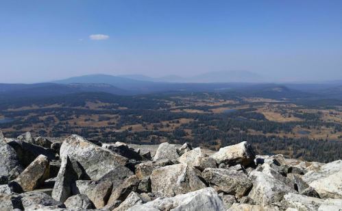 A panoramic view from a rocky summit, showcasing a vast landscape of mountains and valleys under a clear blue sky.