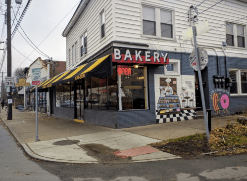 A cozy bakery with a striped awning, large windows, and a colorful mural, located on a street corner.