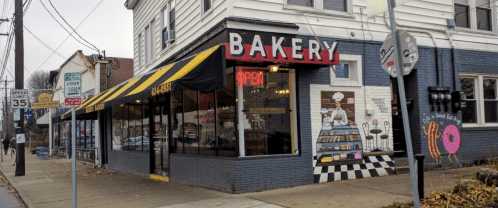 A bakery with a striped awning and "Open" sign, featuring a mural of baked goods on the side wall.