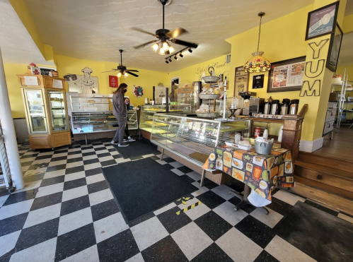 A cozy bakery interior with yellow walls, checkered floor, display cases, and a customer browsing.