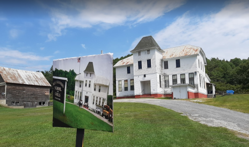 Historic schoolhouse with a sign displaying a vintage photo, set against a green landscape and blue sky.