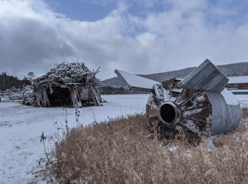 Snow-covered landscape featuring a large metal structure and a wooden pile in the background, with cloudy skies above.