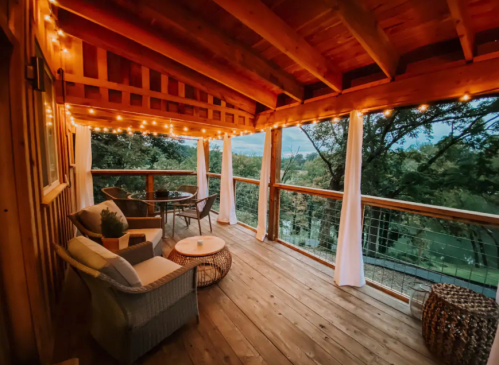 Cozy wooden porch with string lights, seating, and a view of trees and landscape at dusk.