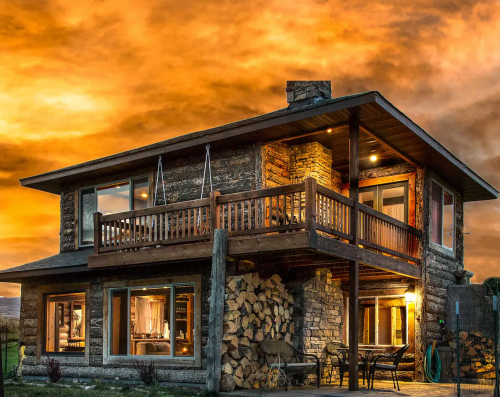 A cozy wooden house with a balcony, surrounded by stacked firewood, under a dramatic orange sunset sky.