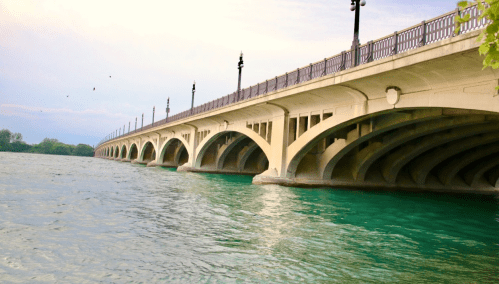 A concrete bridge with arches spans over clear blue water, lined with lampposts under a cloudy sky.