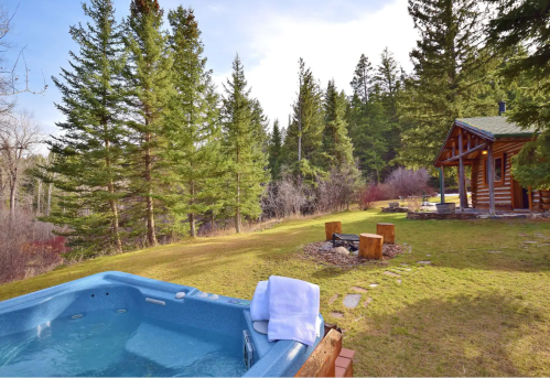 A hot tub beside a grassy area with a fire pit, surrounded by tall trees and a cozy cabin in the background.
