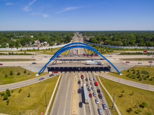 Aerial view of a highway with a blue arch bridge overhead, surrounded by green trees and clear blue skies.