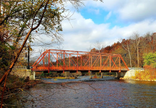 A red truss bridge spans a river, surrounded by autumn foliage and a partly cloudy sky.