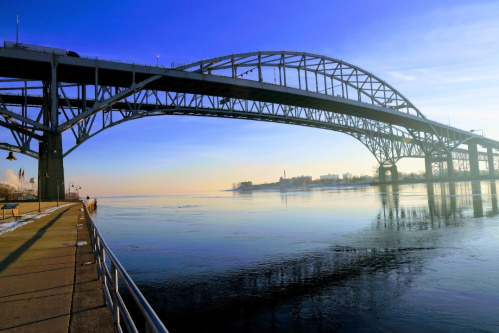 A large steel bridge spans over a calm river, reflecting the blue sky and surrounding landscape at sunrise.