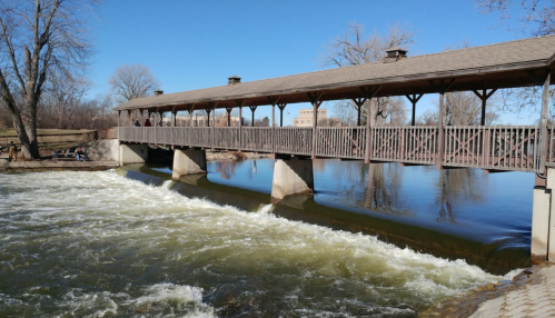 A wooden bridge spans a river with flowing water, surrounded by trees and a clear blue sky.