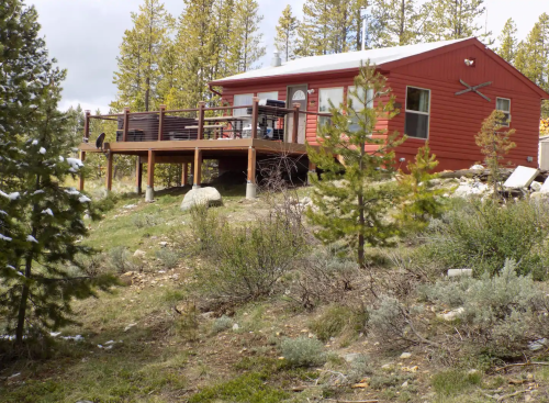 A red cabin with a deck surrounded by trees and shrubs in a natural setting. Snow is visible on the roof.