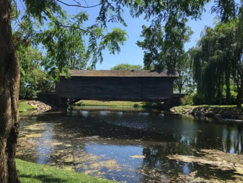 A wooden covered bridge spans a calm pond, surrounded by lush greenery and trees under a clear blue sky.