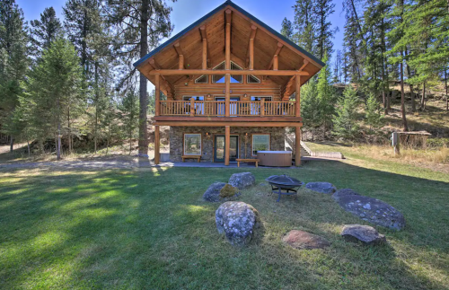 A rustic wooden cabin surrounded by trees, featuring a spacious deck and a fire pit in the grassy yard.