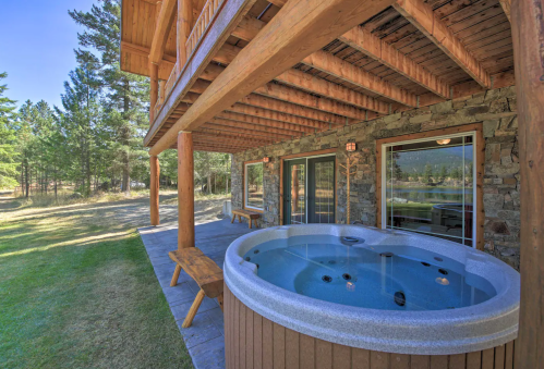 A wooden deck with a hot tub, surrounded by trees and a grassy area, beneath a spacious cabin.