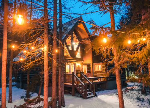 A cozy cabin surrounded by trees, illuminated by string lights, with snow on the ground at dusk.