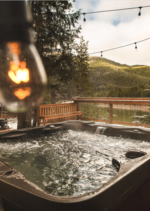 A hot tub with bubbling water, surrounded by trees and mountains, illuminated by a hanging light bulb.