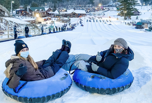 Two people sitting on snow tubes, giving thumbs up, with a snowy hill and a ski resort in the background.