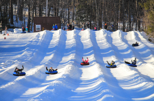 A snowy hill with people tubing down, waving and enjoying a winter day at a tubing park. Trees in the background.
