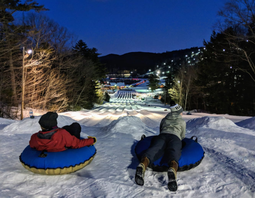 Two people sit on snow tubes at night, overlooking a snowy slope with lights and trees in the background.