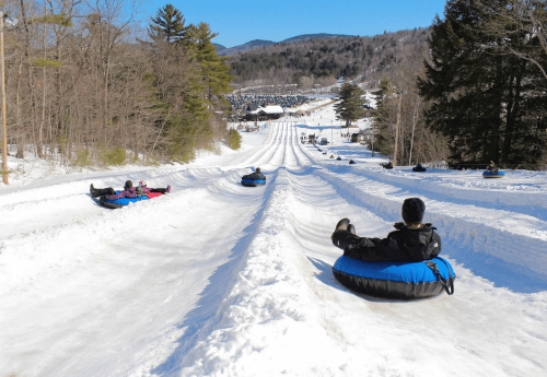 A snowy hill with people tubing down, surrounded by trees and a clear blue sky.