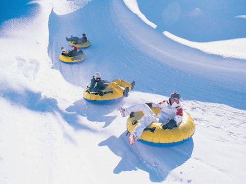 Four people in yellow snow tubes sliding down a snowy slope on a sunny day.