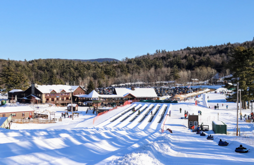 A snowy landscape featuring a ski resort with tubing lanes, buildings, and a parking lot surrounded by trees.