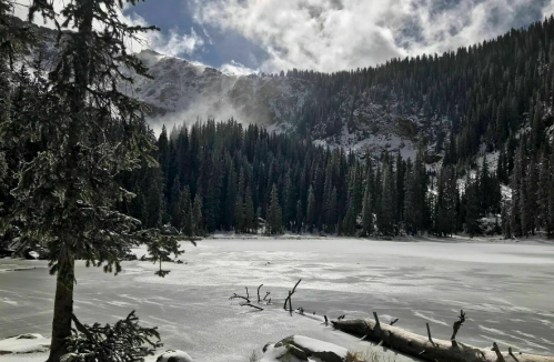 A serene winter landscape featuring a frozen lake surrounded by snow-covered trees and mountains under a cloudy sky.