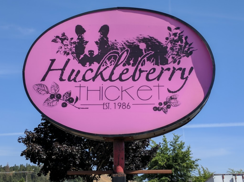 A pink sign for "Huckleberry Thicket," featuring huckleberries and established in 1986, set against a blue sky.